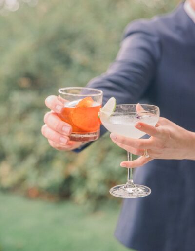 Two hands holding cocktails; one with an orange slice in a tumbler, the other with a lime wedge in a coupe glass. Background is blurred greenery.