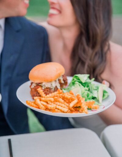 A person holds a plate with a burger, crinkle-cut fries, and a green salad. Two people are smiling in the background.