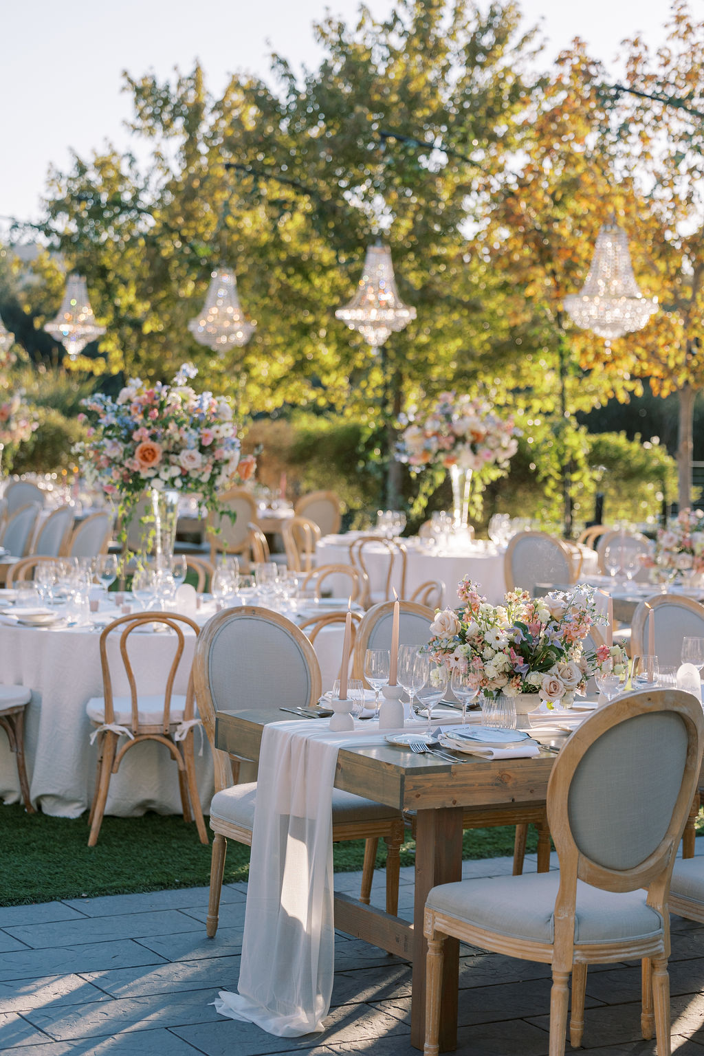 An elegant outdoor wedding reception setup with decorated tables, floral centerpieces, wooden chairs, and hanging chandeliers against a backdrop of trees.