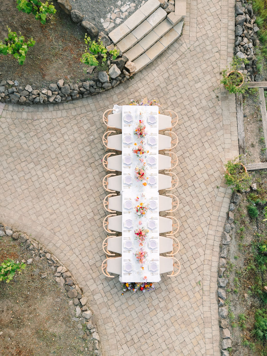 Aerial view of a long outdoor dining table set for twelve with white cloth, plates, and colorful floral centerpieces, positioned on a paved pathway surrounded by greenery.