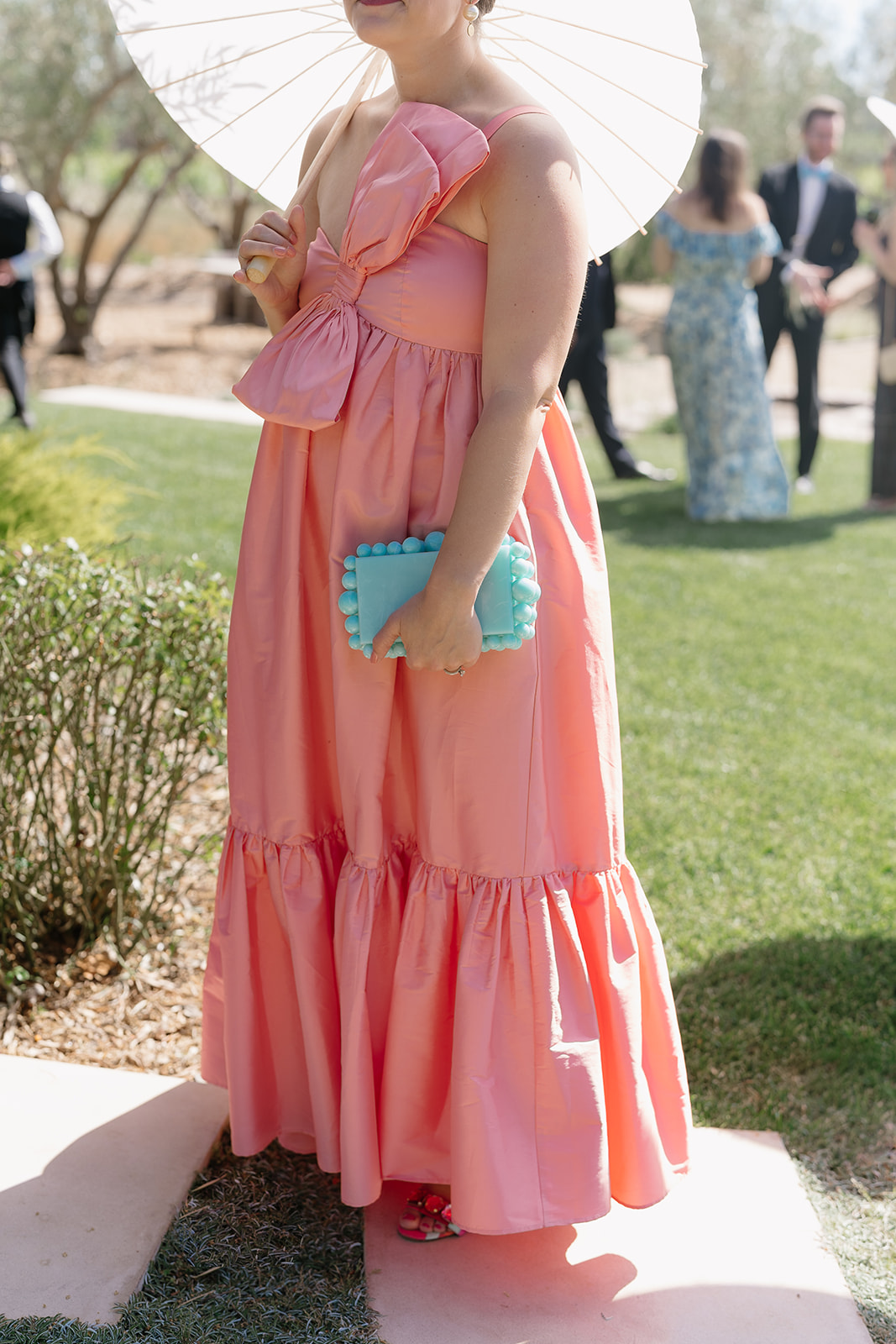 Person in a pink dress holding a blue clutch stands outdoors under a white parasol, with people in the background.