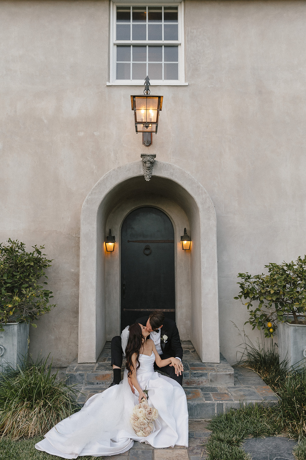 A couple in wedding attire sit on stone steps in front of an arched doorway, surrounded by potted plants. The bride holds a bouquet.