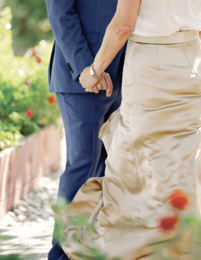 A couple holds hands outdoors. One wears a blue suit, the other a beige dress. The scene is framed by greenery and small red flowers.