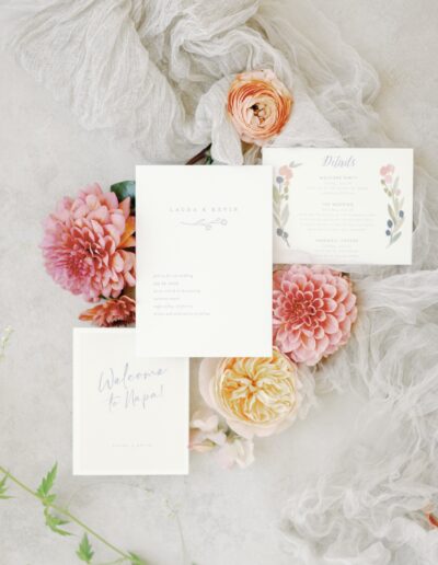 Elegant wedding invitations and details card displayed with pink and yellow flowers on a soft fabric background.