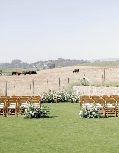 A wedding ceremony setup with wooden chairs and floral arrangements on a green lawn, with cows grazing in a distant field.