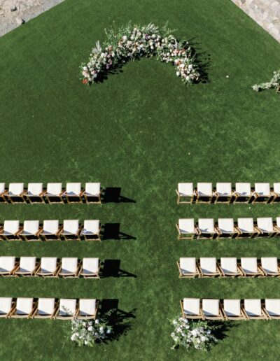 Aerial view of a wedding ceremony setup on a green lawn with rows of white chairs and a floral arch at the front.