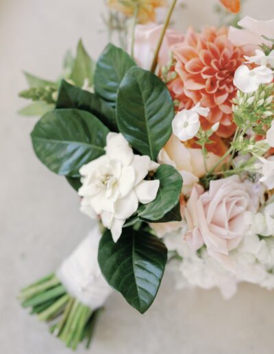 A bouquet with white gardenias, pink roses, orange dahlias, green leaves, and assorted flowers wrapped in white ribbon.