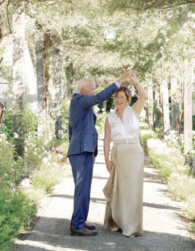 An elderly man in a blue suit and a woman in a white blouse and beige skirt dance together in a garden pathway.