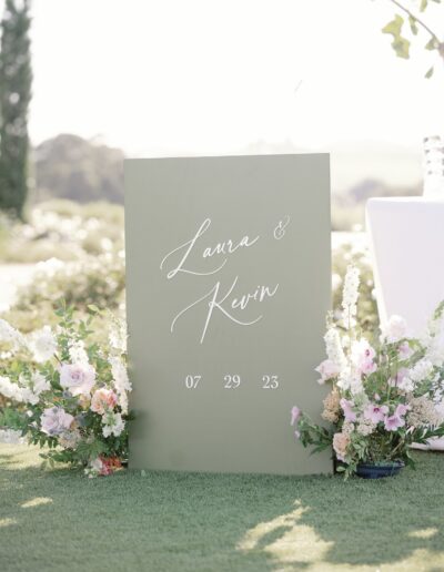 A green wedding sign reads "Laura & Kevin" with the date "07 29 23," surrounded by flowers.