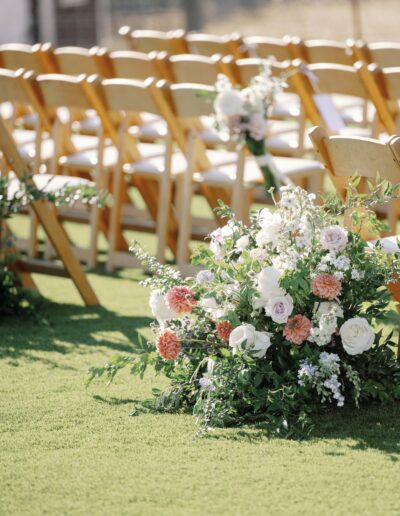 Wooden chairs are arranged in rows on a grassy area. Floral arrangements with white and pink roses decorate the aisle.