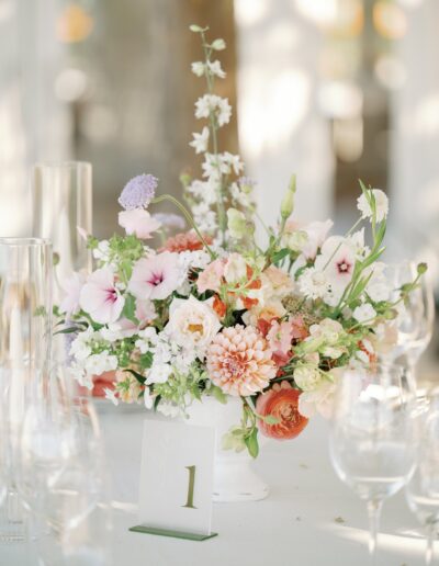 A floral centerpiece with pink, white, and peach flowers in a vase, placed on a table, with clear glassware and a table number card labeled "1".