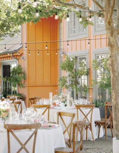 Outdoor dining area with tables set for an event. Round tables with white tablecloths and wooden chairs, under string lights, next to an orange building.