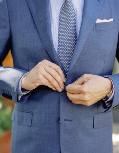 A person wearing a blue suit and patterned tie adjusts their jacket button.