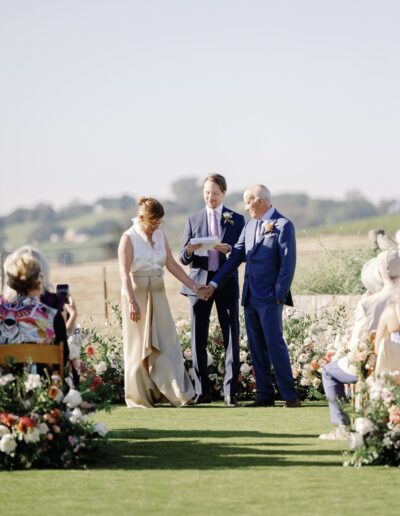 Two people are standing, holding hands, facing each other at an outdoor wedding ceremony. An officiant is present, with flowers and chairs nearby.