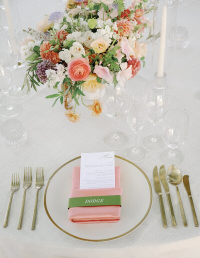 A table setting with a floral centerpiece, menu on a pink napkin, and gold cutlery. The napkin has a green band with the word "Dodge.