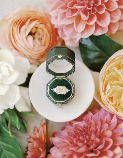 An ornate ring with a square gemstone in an open green box, surrounded by pink and white flowers on a white surface.