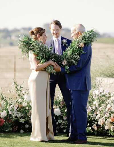 A couple stands facing each other outdoors, exchanging large green garlands, with another person standing in the background. There are flowers and greenery around them.