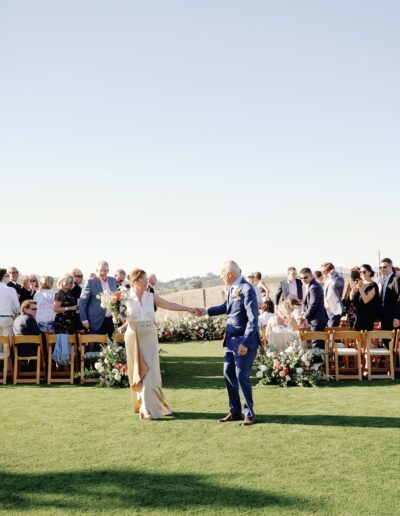 A couple holding hands prepares to walk down a grassy aisle, surrounded by seated guests and floral arrangements at an outdoor event.