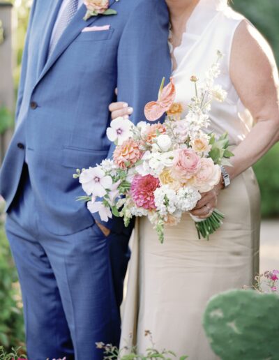 A couple in formal attire embrace, with the woman holding a colorful bouquet of flowers, standing outdoors.