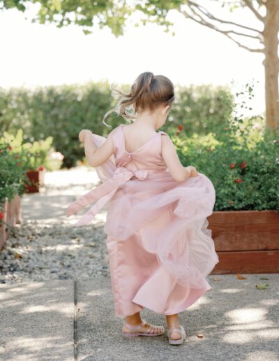 A young girl in a pink dress walks along a garden path surrounded by greenery and sunlight.