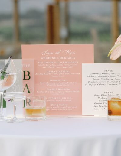 Wedding bar menu on a table with various cocktails, flowers, and a view of a landscape in the background.