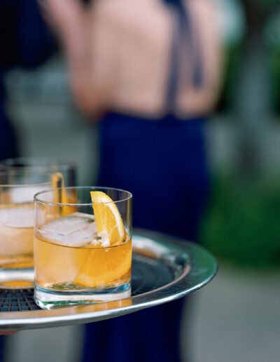 A tray with four glasses of iced cocktails, each garnished with an orange slice, is held outdoors at a Park Winters wedding. People are blurred in the background.