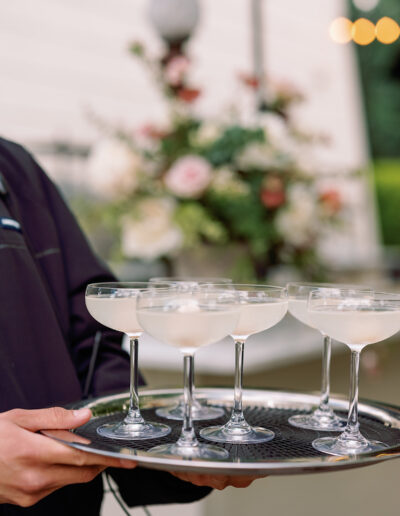 Close-up of a server at Park Winters Weddings, gracefully holding a tray with five champagne-filled glasses, set against a blurred backdrop of flowers and lush greenery.