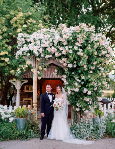 The bride and groom stand under a floral archway at Park Winters, dressed in formal wedding attire, surrounded by lush greenery and a charming picket fence.