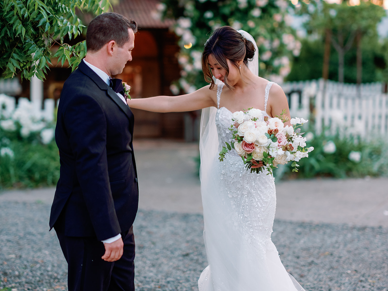 A bride in a white gown holding a bouquet stands beside a groom in a black suit outdoors, surrounded by lush greenery and flowers—a picture-perfect moment at Park Winters Weddings.