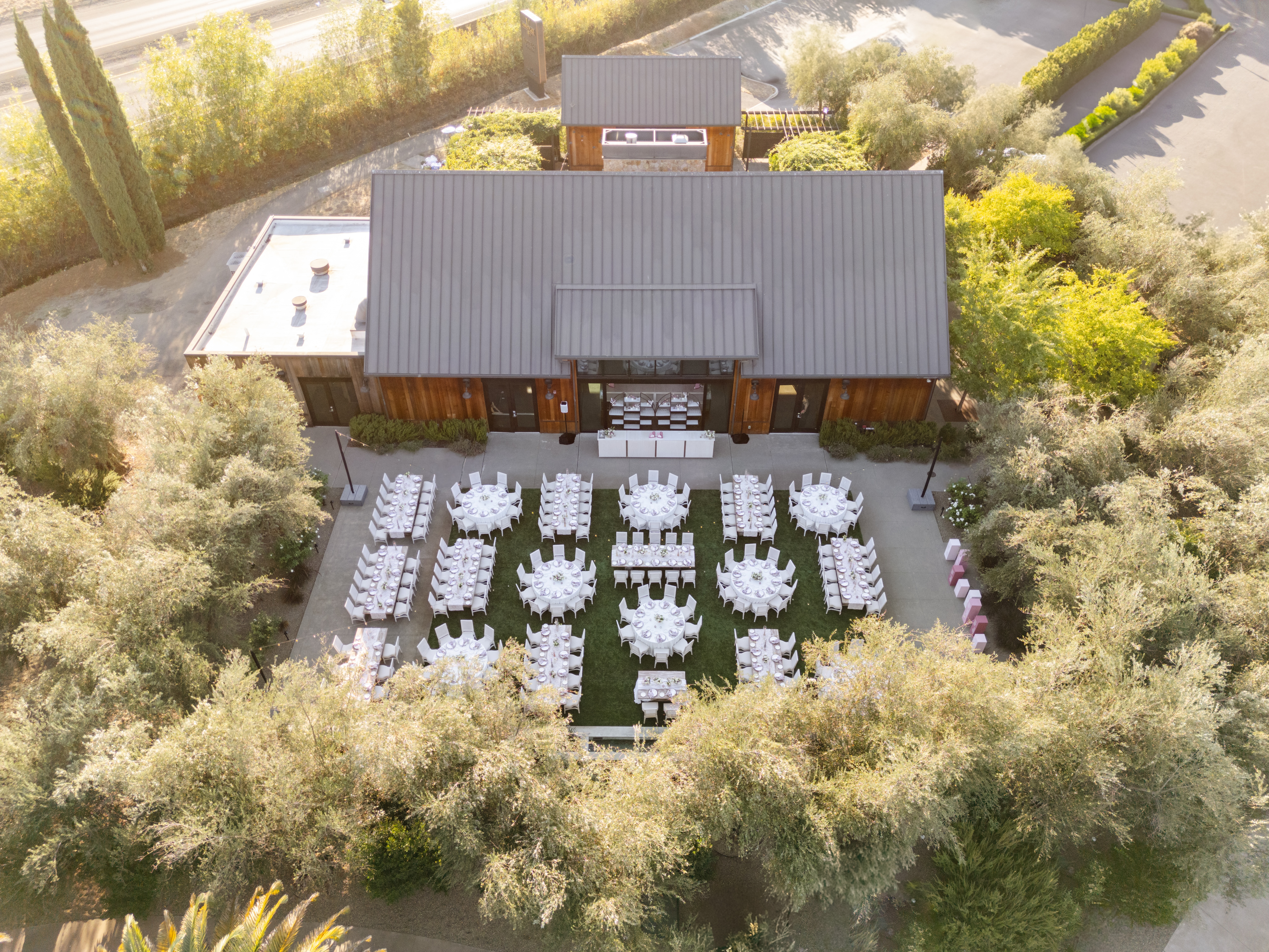 Outdoor wedding setup with white tables and chairs arranged symmetrically in a garden setting, surrounded by trees, adjacent to a wooden building.