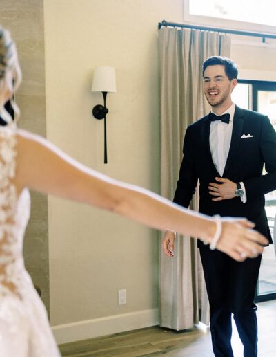 A bride and groom share a happy moment indoors. The bride is in a white dress with lace details, and the groom is in a black tuxedo. They are facing each other with outstretched arms.