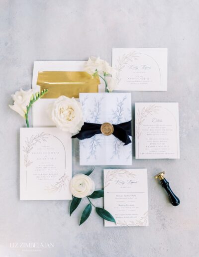 Wedding invitation suite with floral designs, featuring a sealed letter with a black ribbon, surrounded by white flowers and greenery.