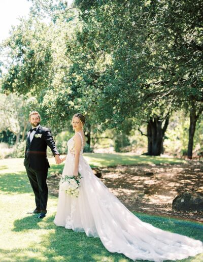 A bride and groom stand hand in hand on a grassy area under trees. The bride wears a long white dress with a train, and the groom is in a black suit.