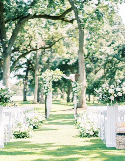 Outdoor wedding ceremony setup with white chairs, floral arrangements on pedestals, and a flower-adorned archway under tall trees.