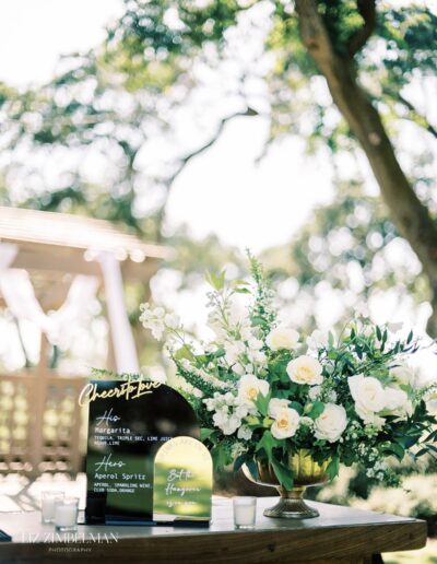 Outdoor wedding setup with a drink menu sign, floral arrangement of white roses, greenery, and candles on a wooden table. A pergola and trees are in the background.