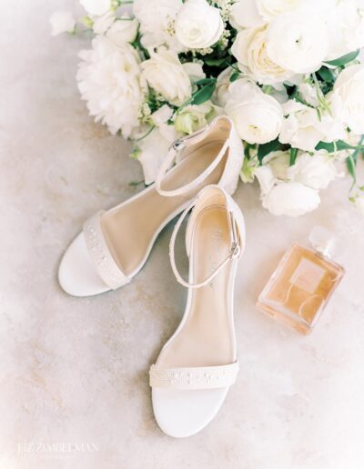 White open-toe bridal shoes with ankle straps on a textured surface, surrounded by white flowers and a bottle of perfume.