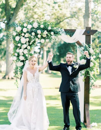 Bride and groom celebrate under a flower-adorned wooden arch outdoors, both smiling and raising their hands in joy.