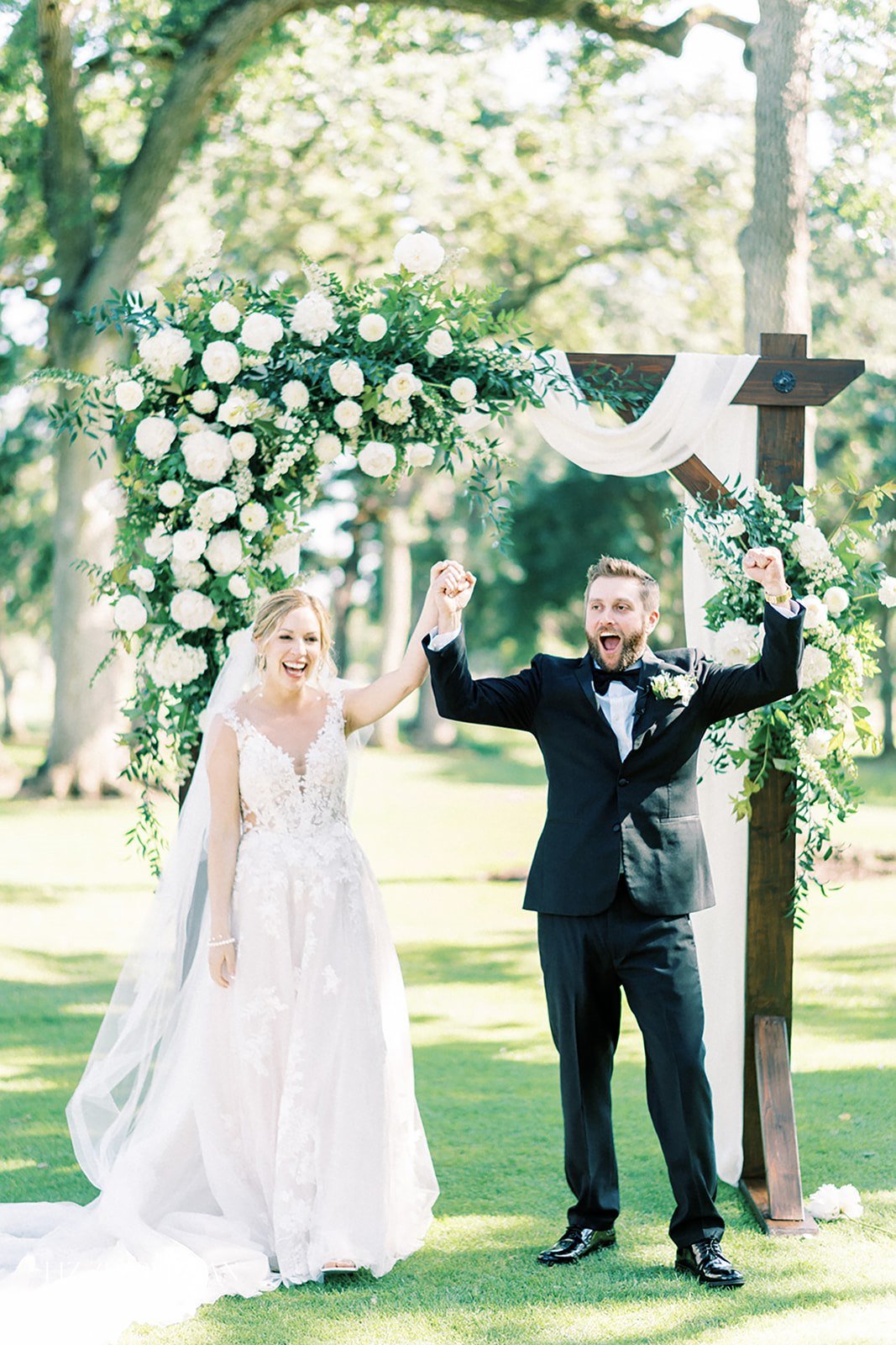 Bride and groom celebrate under a flower-adorned wooden arch outdoors, both smiling and raising their hands in joy.