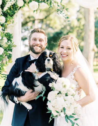 A couple in wedding attire poses outdoors, smiling and holding a black and white dog. Flowers and greenery embellish the scene.