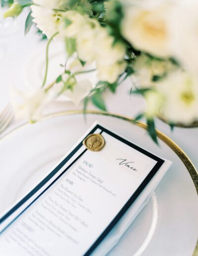 A close-up of a wedding menu on a plate. The menu features a gold wax seal and lists options for salad and main courses. White flowers are slightly blurred in the background.