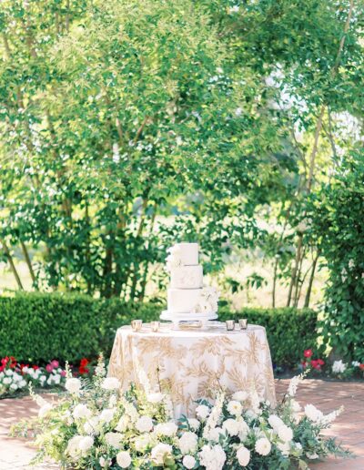 A tiered white cake on a decorated table with floral arrangements in an outdoor setting.