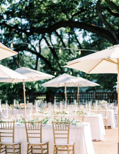 An outdoor event setup with white tablecloths, gold chairs, and white umbrellas under trees. String lights are visible above the tables.