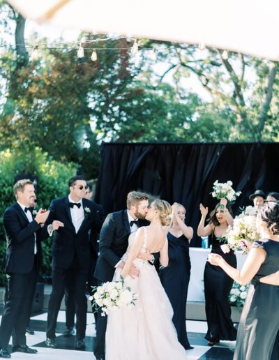 A couple shares a kiss at an outdoor wedding ceremony, surrounded by a cheering bridal party dressed in black formal attire.