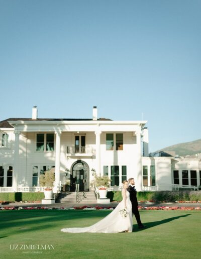 A bride and groom walk hand in hand on a manicured lawn in front of a large white mansion.