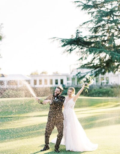 A couple in wedding attire celebrates outdoors by spraying champagne, with trees and a building in the background.