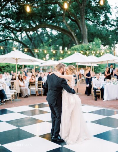 A couple dances on a checkered floor at an outdoor wedding reception with guests seated and string lights above.