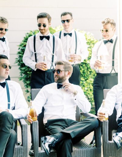 A group of men in sunglasses and formal attire, with bow ties and suspenders, sit and stand around outdoors, holding bottled drinks and engaging in conversation.