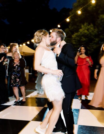 A couple dancing closely on a checkered floor under string lights, surrounded by people in formal attire.