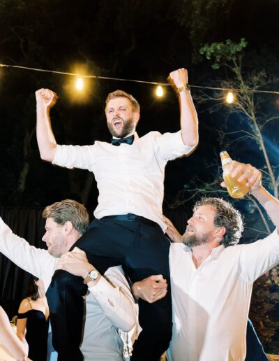 A man in a white shirt with his arms raised is lifted on the shoulders of two others at a nighttime outdoor event. One person holds a drink. String lights are visible in the background.