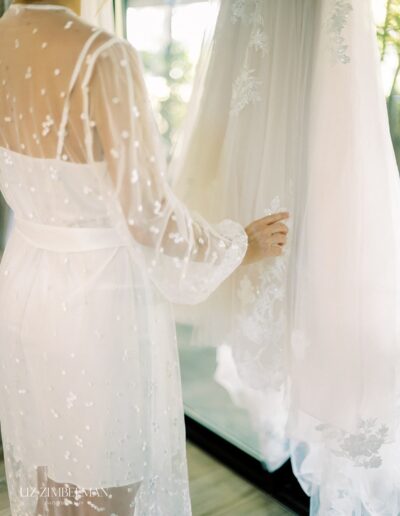 A person in a sheer white robe touches a hanging white lace dress in a softly lit room.